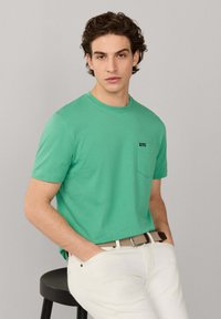 Jeune homme aux cheveux bouclés foncés porte une chemise verte à manches courtes et un pantalon blanc, assis sur un tabouret noir devant un fond uni.