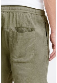 Olive green shorts with a crinkled texture, elastic waistband, and a side pocket visible on the back, showcasing functional design elements.