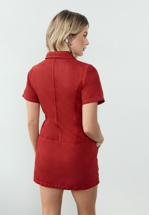 Femeie cu păr scurt și blond, purtând căști wireless albe și o rochie roșie cu mâneci scurte și buzunare, stând cu mâinile în buzunare, orientată lateral.