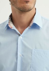 Chemise en coton bleu clair avec un col à motifs beige, rouge et noir. Dotée d'une poche sur le devant et d'une fermeture à boutons blancs.
