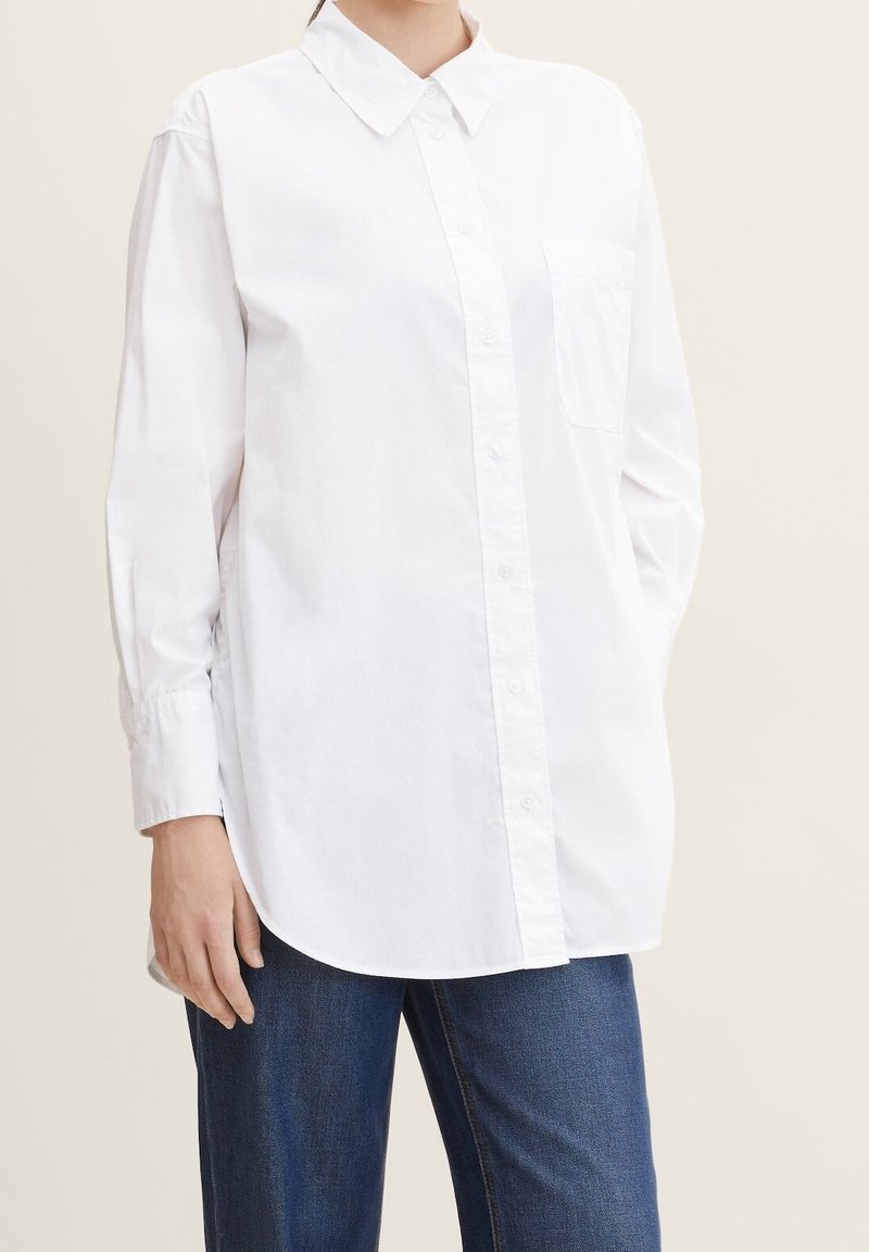 Personne portant une chemise blanche à manches longues avec une poche poitrine et un jean bleu, debout sur un fond clair uni.
