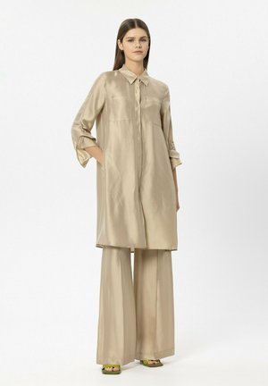 Woman wearing beige long satin shirt with pockets and wide-leg pants, standing with hands in pockets, on white background.