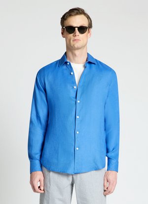Homme portant des lunettes de soleil noires, une chemise bleue brillante boutonnée sur un t-shirt blanc et un pantalon gris clair à rayures sur fond uni.