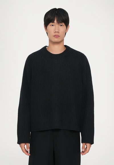 Un giovane con capelli neri e lisci, che indossa un maglione a coste nero e pantaloni neri, in piedi di fronte a uno sfondo bianco uniforme.