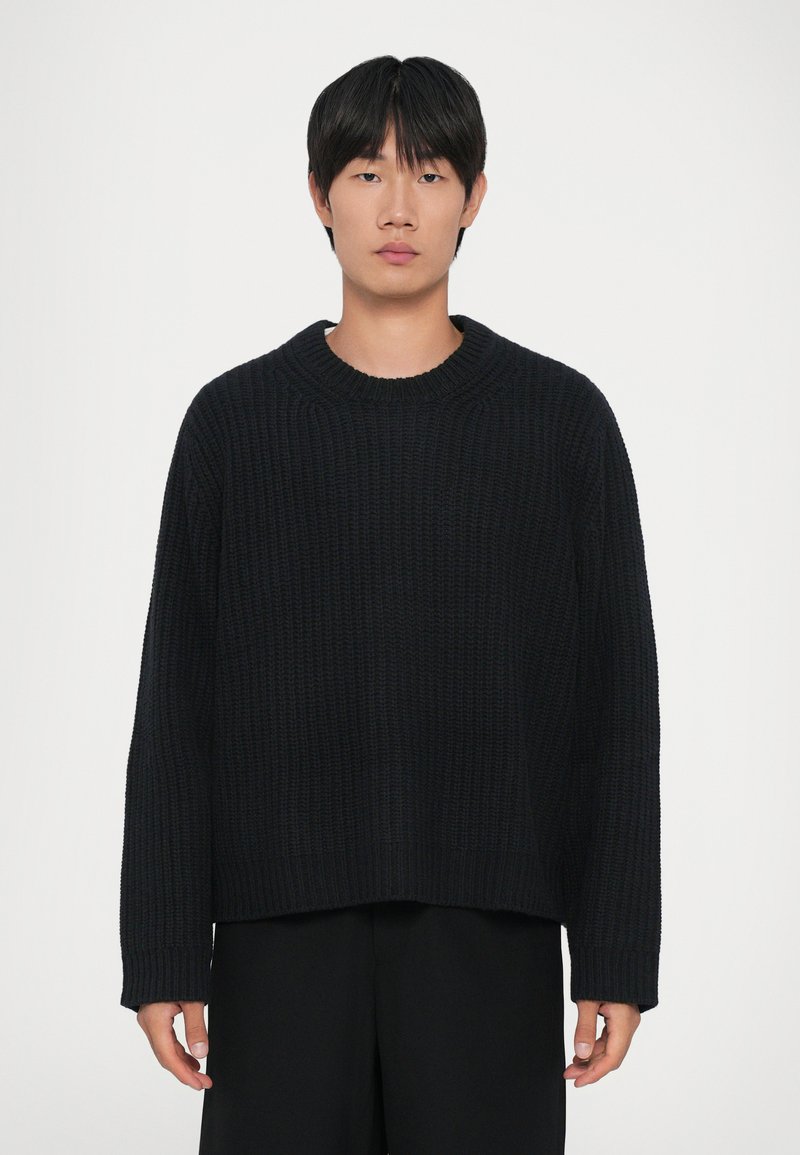 Un giovane con capelli neri e lisci, che indossa un maglione a coste nero e pantaloni neri, in piedi di fronte a uno sfondo bianco uniforme.