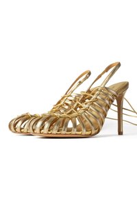 Sandale cu toc aurii metalice, cu curea încrucișată, design cu vârf deschis și tocuri subțiri. Textură netedă și detalii minime.