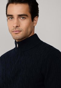 Maglione blu navy a lavorazione a trecce con colletto alto e dettaglio zip argentata. La texture è a coste, aggiungendo profondità al design.
