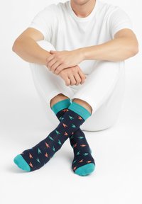 Osoba sedící se zkříženýma nohama, obutá v tmavě modrých ponožkách s oranžovými a tyrkysovými vzory ptáků, v kombinaci s bílými kalhotami a košilí.