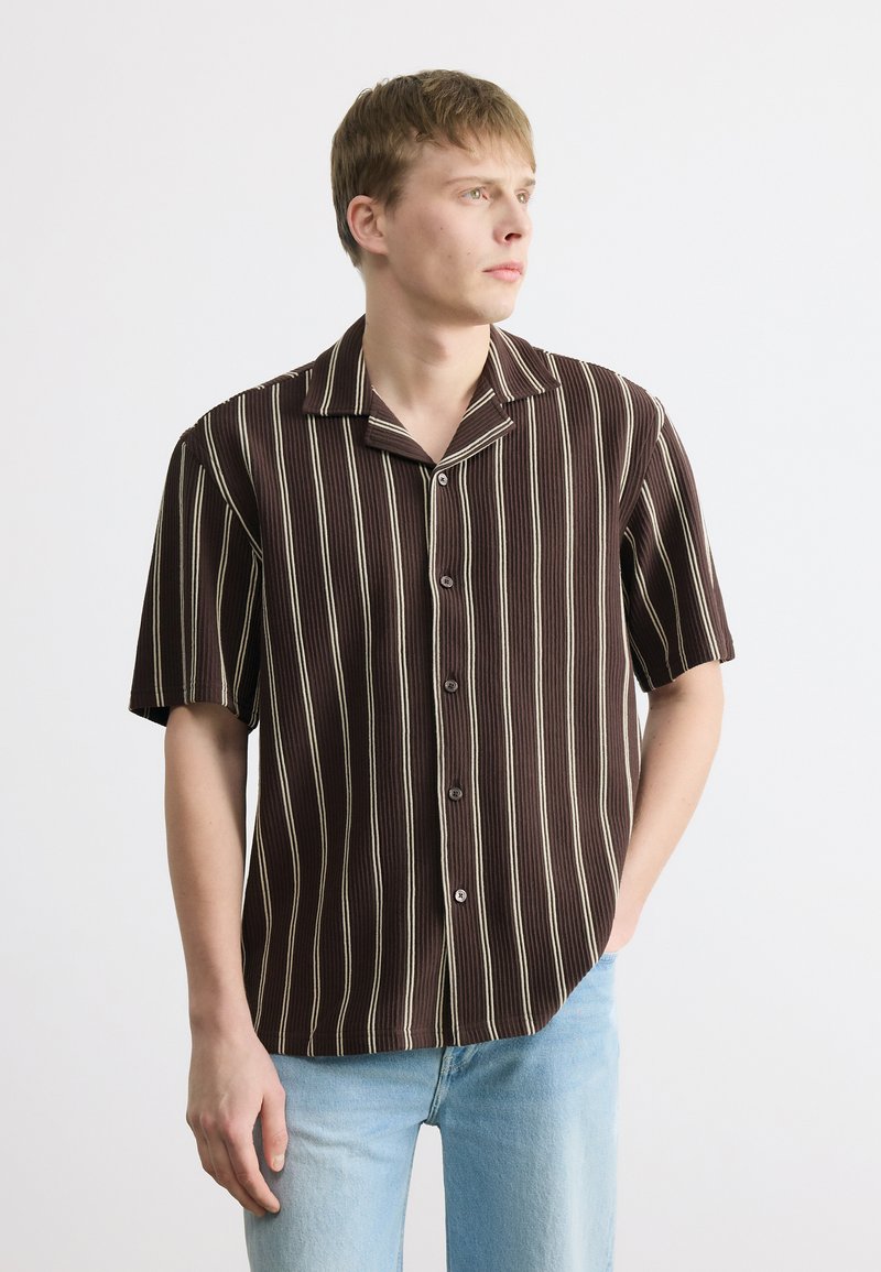 Νεαρός άνδρας με κοντά μαλλιά που φορά καφέ κοντομάνικο πουκάμισο με κάθετες μπεζ ρίγες και ανοιχτόχρωμα μπλε τζιν, κοιτάζει προς τα δεξιά.