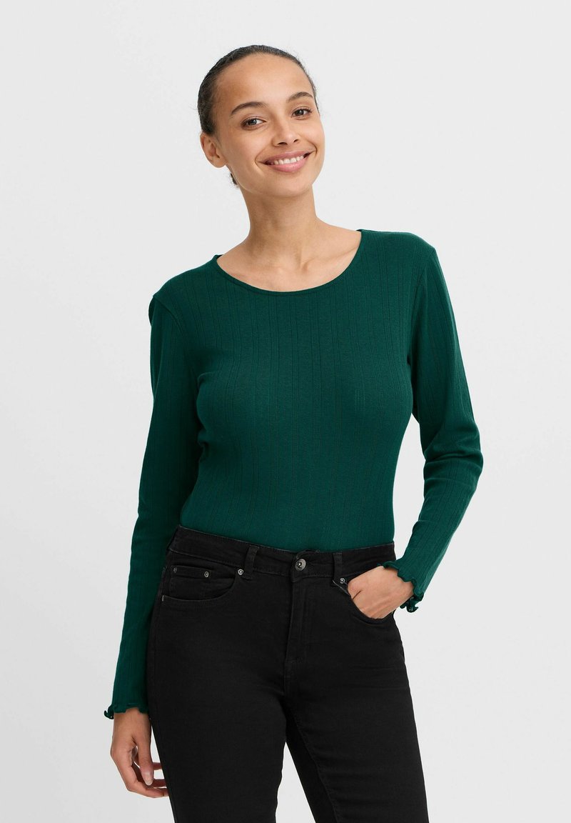 Grünes, geripptes Langarmoberteil mit rundem Ausschnitt, kombiniert mit hochgeschnittenen schwarzen Jeans, die dezente Akzente und Taschendetails aufweisen.