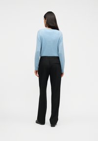 Γυναίκα με πλάτη, φοράει ανοιχτό μπλε πουλόβερ, μαύρο παντελόνι φαρδύ στην πόδι και μαύρα παπούτσια πάνω σε απλό λευκό φόντο.