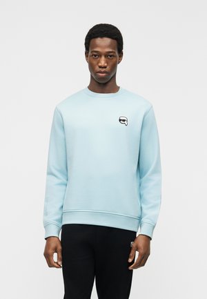 Mann trägt hellblauen Pullover mit kleinem schwarz-weißem Emoji-Logo auf der Brust und schwarze Hose, steht vor weißem Hintergrund.