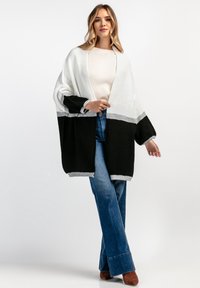 Czarny i biały, oversize'owy sweter z dzianiny o szerokich rękawach, z szarym poziomym paskiem. Nosi się go na jasnej bluzce i niebieskich dżinsach.