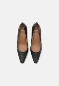 Scarpe con tacco alto in pelle nera con design a punta. L'interno è di colore beige con branding. Texture liscia, stile minimalista senza accenti visibili.
