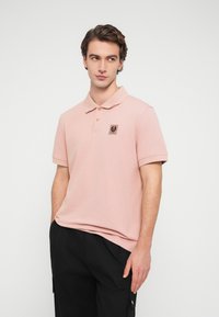 Jeune homme portant un polo rose clair avec un logo en écusson sur la poitrine et un pantalon noir, debout devant un fond blanc uni.