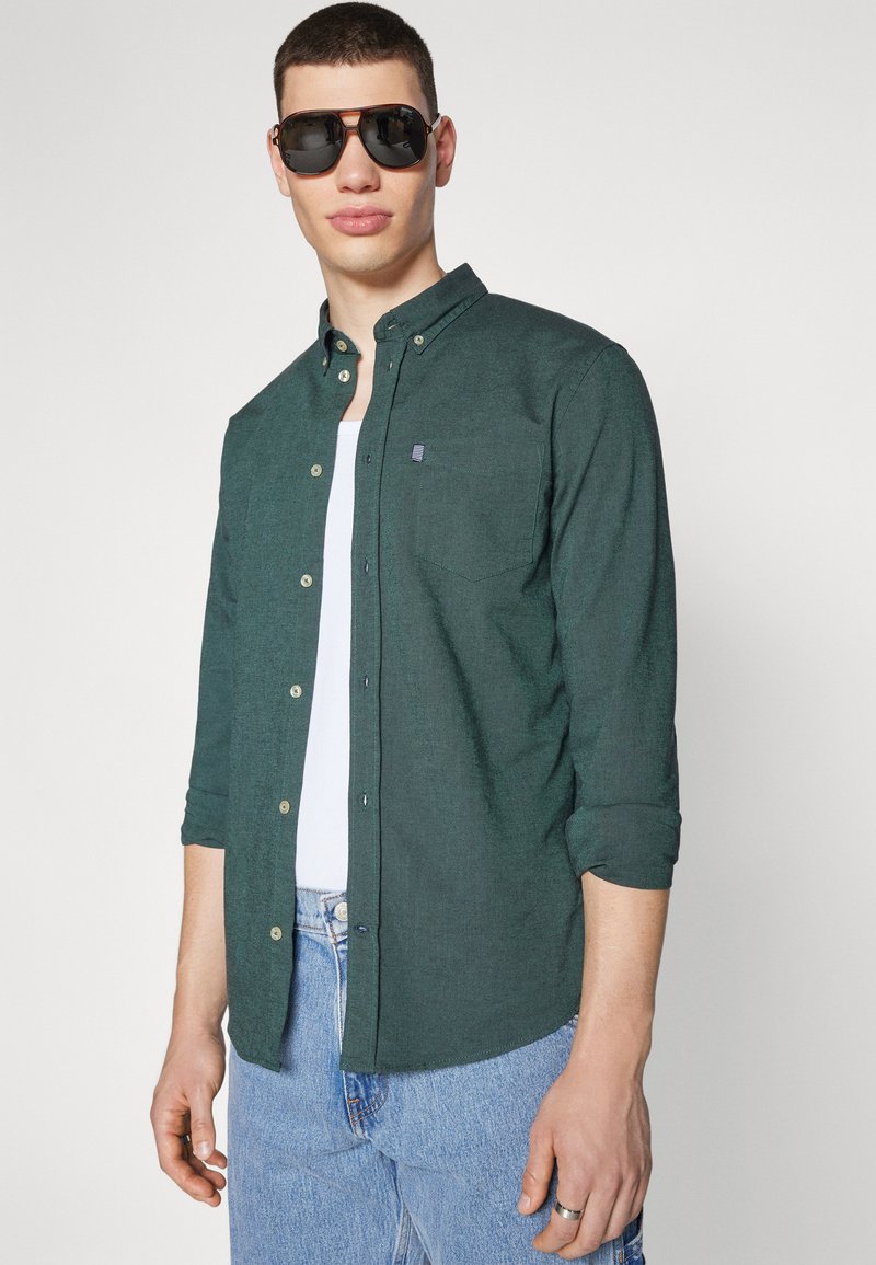 Camisa de botones de color verde oscuro en una tela texturizada, con un bolsillo en el pecho y botones de color claro, llevada sobre una camiseta sin mangas blanca.