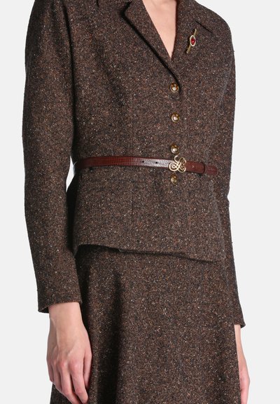 Femme portant un blazer en tweed marron boutonné avec une jupe assortie, une fine ceinture marron avec boucle dorée et une broche dorée avec une pierre rouge sur le revers.
