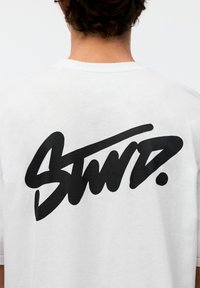 Hvid bomulds t-shirt med stor sort grafisk tekst "Stwd." på ryggen. Korte ærmer og afslappet pasform.