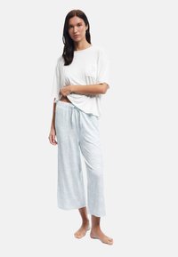 Vrouw in witte losse t-shirt en lichtblauwe bloemen baggy broek, staand op blote voeten tegen een strakke witte achtergrond.