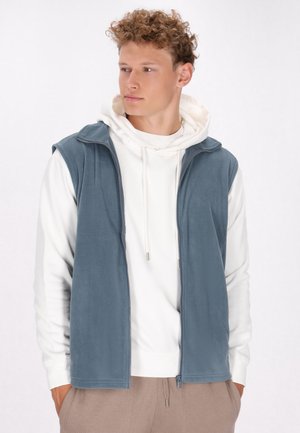 Blauer Fleece-Weste mit Reißverschluss auf der Vorderseite, die über einem weißen Hoodie getragen wird. Glatte Textur, ärmelloses Design und kontrastierende helle Nähte.