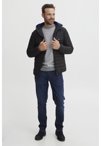 Svart puffjakke med blå hette, glidelås i front og synlig brystlomme, kombinert med grå genser og mørkeblå jeans. Svarte støvler.
