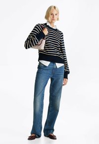 Maglione a strisce blu navy e crema, jeans blu e ballerine bordeaux. La modella tiene una borsa a motivi con manico superiore. Camicia bianca sotto il maglione.