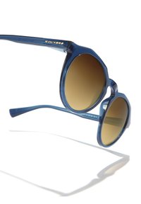 Modré osmihranné sluneční brýle s hnědými šedými skly s gradientem, průhlednými obroučkami a zlatým logem na stranici. Hladká textura a lehký design.
