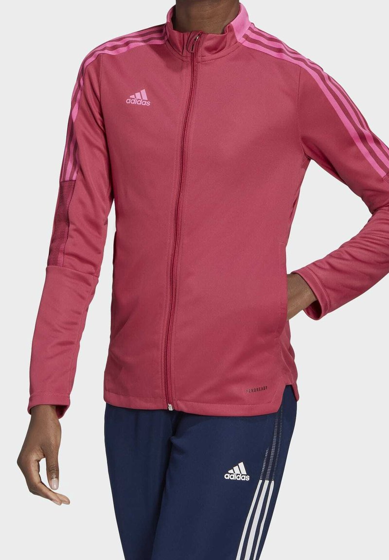 Chaqueta deportiva rosa con cuello alto, cremallera frontal y tres rayas rosas en cada hombro. Presenta una manga texturizada y un logo.