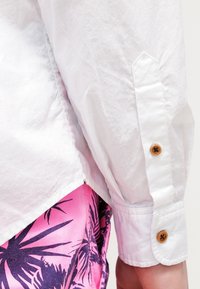 Vit, rynkad skjorta med långa ärmar och manschetter med knapp, kombinerad med rosa shorts med ett marinblått tropiskt bladmönster.