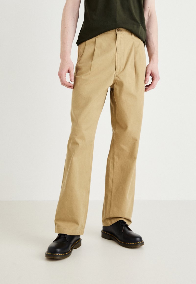 Khaki cotton trousers with a straight-leg design, featuring front pleats and a button closure; paired with black leather shoes.