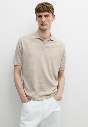 Jeune homme aux cheveux châtain clair portant un polo beige à manches courtes et un pantalon blanc, les mains dans les poches, debout contre un mur uni.