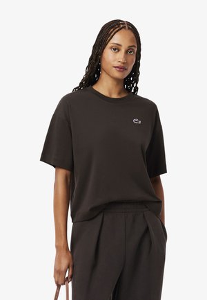 Camiseta de algodón negra con mangas cortas, cuello redondo y un pequeño logotipo de cocodrilo bordado en el pecho. Combinada con pantalones negros.