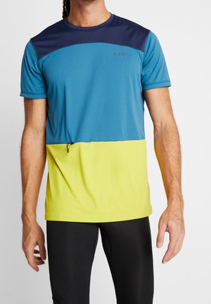 Hombre vistiendo una camiseta deportiva de manga corta con bloques de color en azul marino, azul y amarillo, combinada con mallas negras.