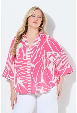 Femme blonde portant de grandes boucles d'oreilles en cercle, une chemise ample à boutons rose et blanche à motifs, et un pantalon blanc, debout devant un fond blanc.