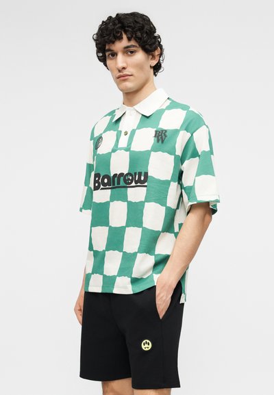 Mladý muž s kučeravými vlasmi nosí zeleno-biele kárované polo tričko s logom "Barrow" a čierne šortky s žltou nášivkou s úsmevom.