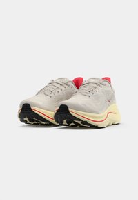 Scarpe da corsa beige chiaro con parte superiore testurizzata, dettagli rossi sul colletto e una suola ammortizzata bianca e gialla con un motivo di trazione nero.