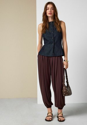 Žena oblečená v tmavom bezrukávovom zapínanom topu, voľných hnedých nohaviciach, čiernych sandáloch, so zlatým náramkom, držiaca hnedú kabelku s bodkami, stojaca v interiéri.