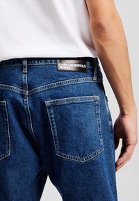 Jean en denim bleu foncé avec une patch logo noir à la taille, dotés de poches arrière standard et de détails de couture visibles.