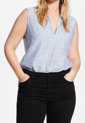 Kobieta w niebiesko-białej bluzce w paski bez rękawów schowanej w wysokich czarnych spodniach z guzikami, ręce w kieszeniach.