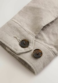 Poignets de chemise en lin beige avec une couture détaillée et deux boutons ronds, marbrés marron foncé, attachés sur un fond blanc.