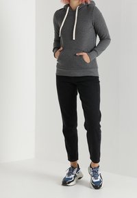 Persona che indossa una felpa grigia scura, pantaloni neri e scarpe da ginnastica blu e bianche, in piedi con le mani nelle tasche della felpa.