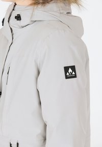 Helle graue Winterjacke mit einer abzipbaren Kapuze, die mit einem Kunstpelzbesatz und einem schwarzen Logo-Patch am linken Ärmel versehen ist. Glattes, wetterbeständiges Material.