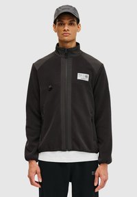 Zwarte fleecejack met ritsluiting, hoge kraag, zijzakken en een label op de borst. Draagt over een wit shirt, met een grijze pet.