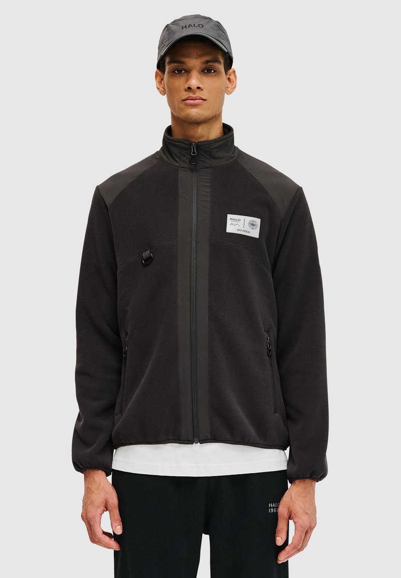 Zwarte fleecejack met ritsluiting, hoge kraag, zijzakken en een label op de borst. Draagt over een wit shirt, met een grijze pet.