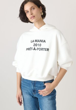 Fehér, nagy méretű kapucnis pulóver elülső zsebbel, fekete "LA MANIA 2010 PRÊT-À-PORTER" felirattal. Kék farmernadrággal párosítva.