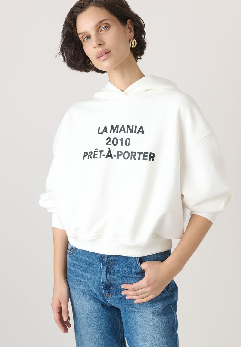 Biała, oversized bluza z kapturem z przednią kieszenią, z czarnym napisem "LA MANIA 2010 PRÊT-À-PORTER". Połączona z niebieskimi dżinsami.