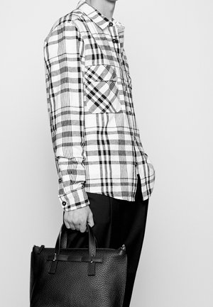 Person iført en sort-hvid ternet langærmet skjorte, der holder en tekstureret sort lædertaske i håndtagene.