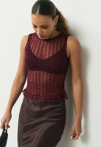 Burgundy textured top with sheer overlay, featuring a frilled hem and sleeveless design, paired with a dark satin skirt.