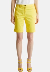 Pantalones cortos de algodón amarillos con un ajuste entallado, que presentan un dobladillo con vuelta y cierre frontal con botones. Combinados con sandalias amarillas a juego con tiras.