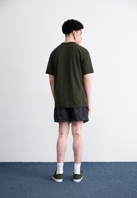 Mørkegrøn kortærmet t-shirt, sorte mønstrede shorts, hvide ankelstrømper, grønne sneakers, set fra ryggen mod en ensfarvet baggrund.